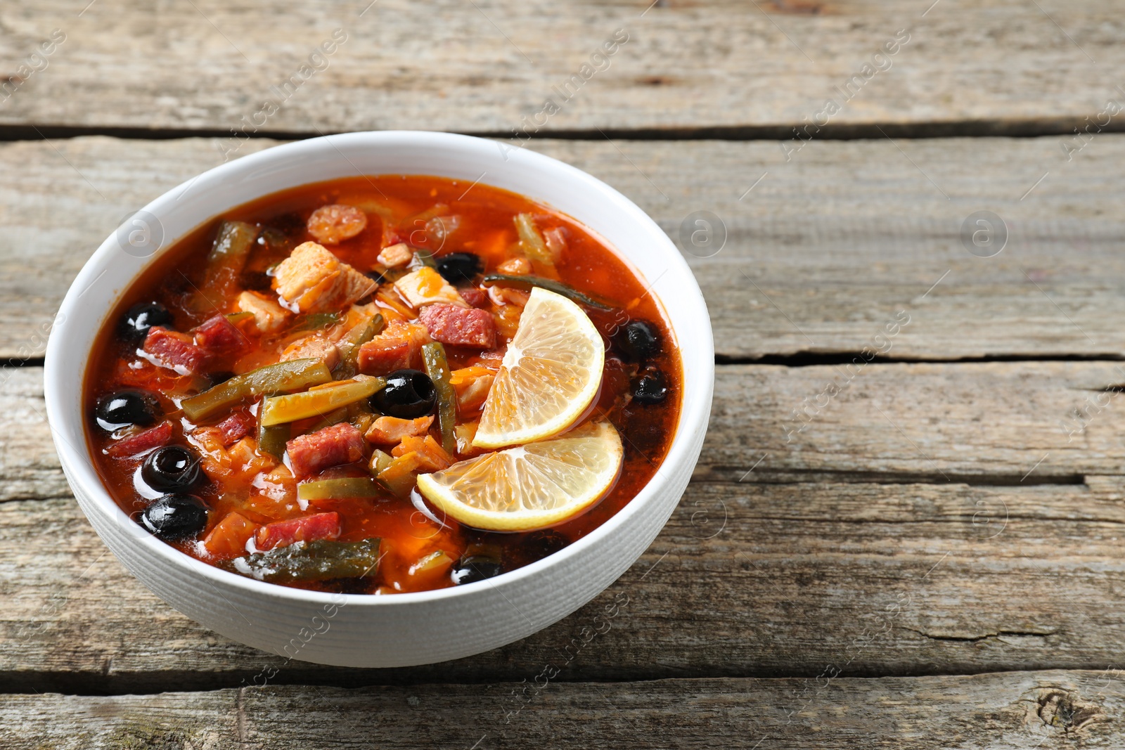 Photo of Meat solyanka soup with sausages, olives and vegetables in bowl on wooden table. Space for text