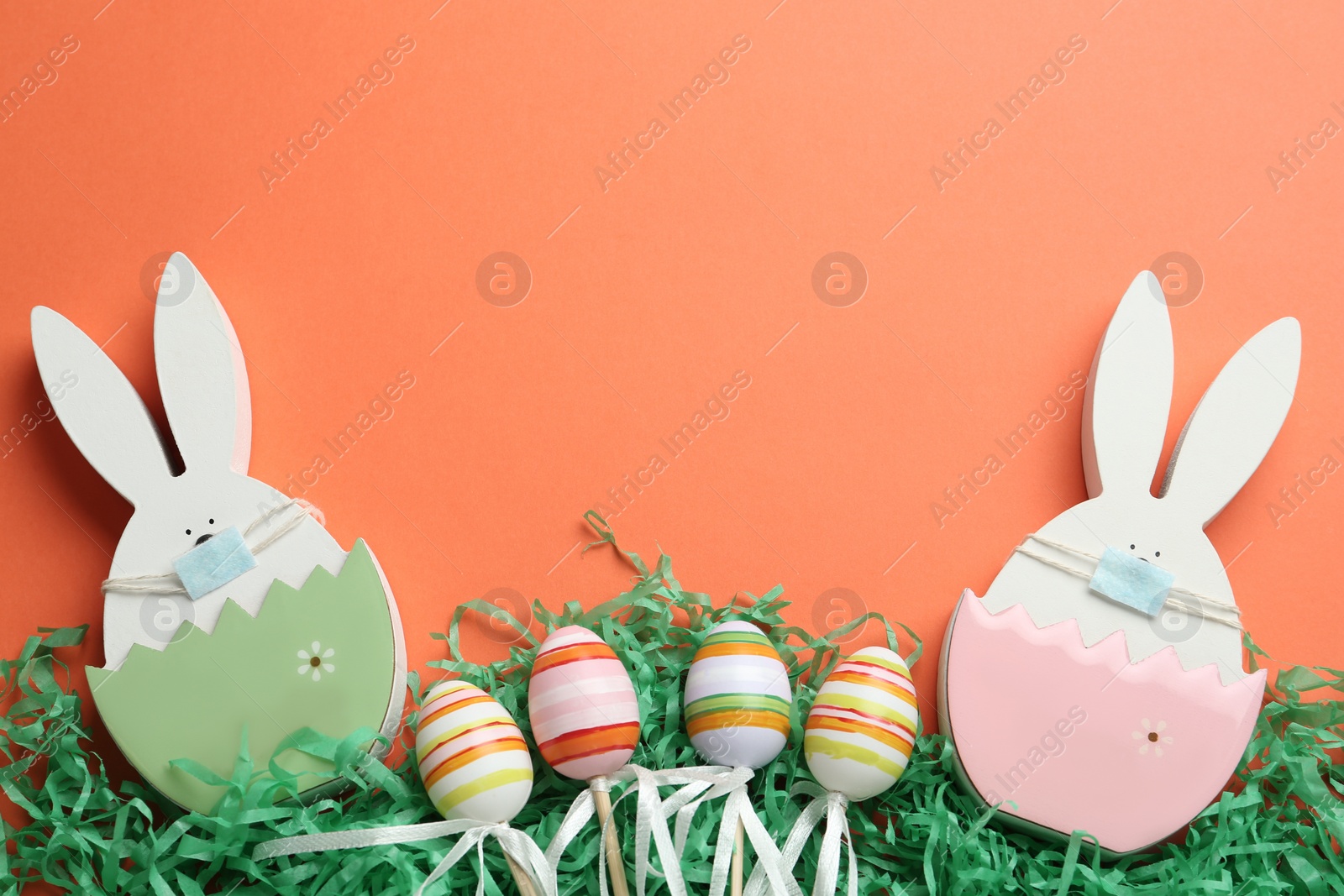 Photo of Wooden bunnies with protective masks, eggs and space for text on coral background, flat lay. Easter holiday during COVID-19 quarantine