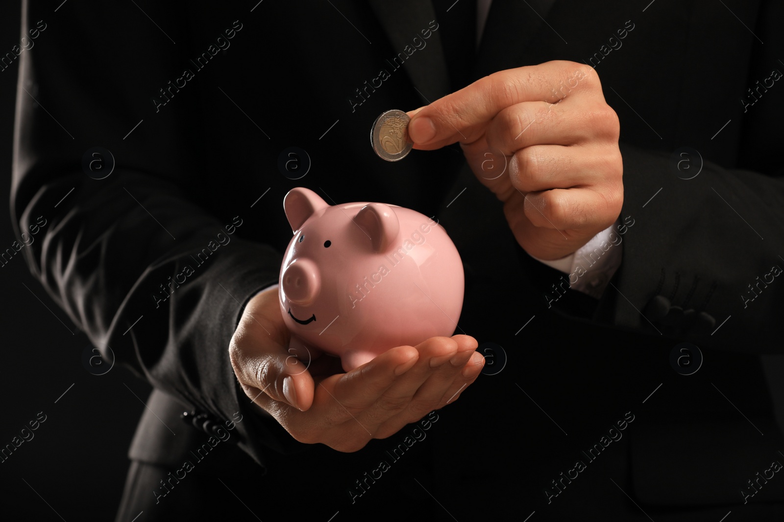 Photo of Businessman putting coin in piggy bank on black background, closeup