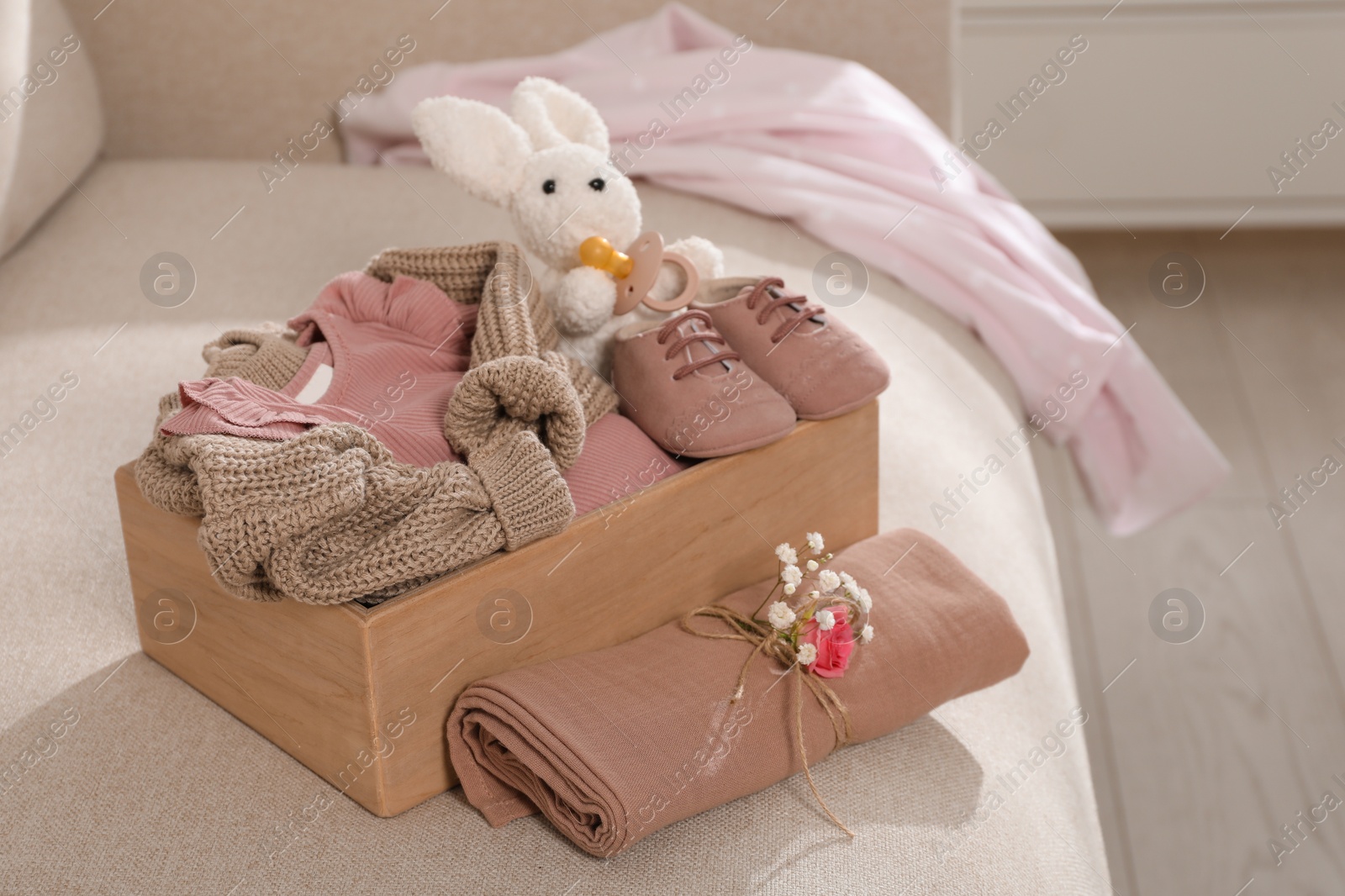 Photo of Wooden crate with children's clothes, shoes, toy bunny and pacifier on sofa in room