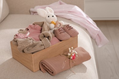 Photo of Wooden crate with children's clothes, shoes, toy bunny and pacifier on sofa in room