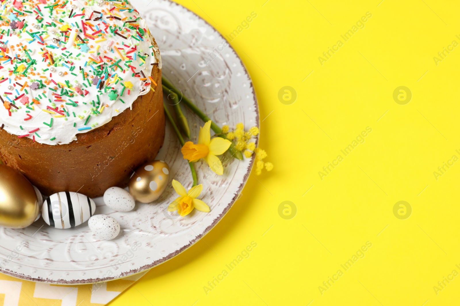 Photo of Traditional Easter cake with sprinkles, painted eggs and beautiful spring flowers on yellow background, space for text