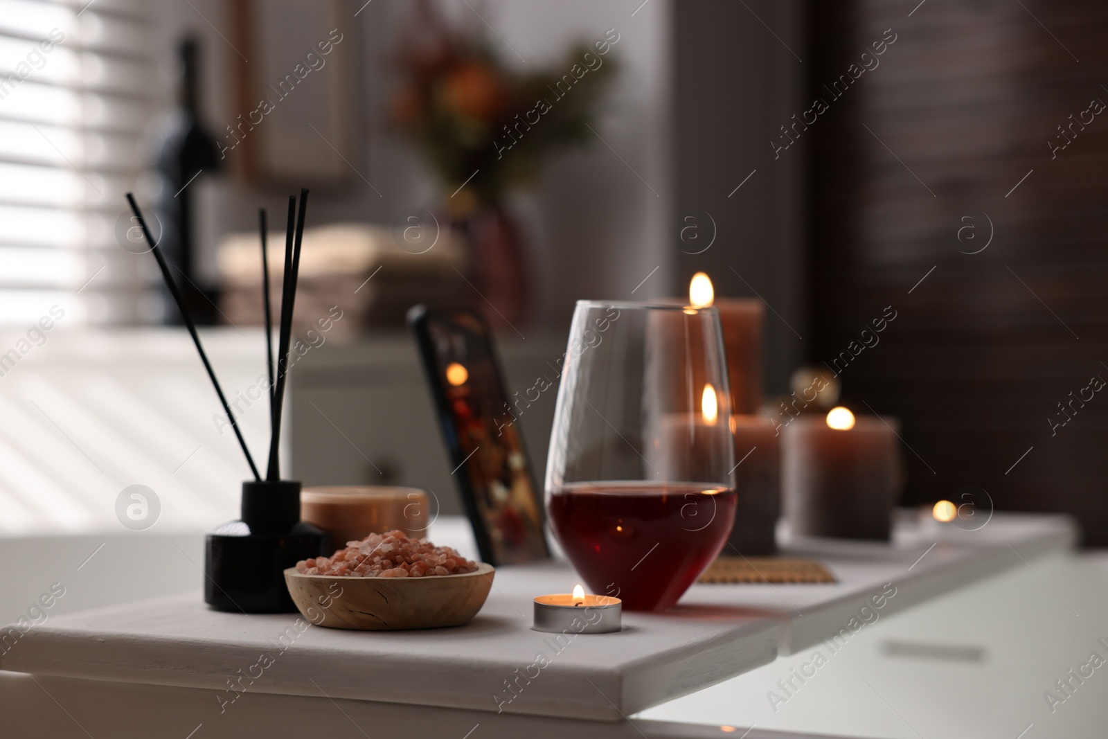 Photo of White wooden tray with glass of wine, beauty products and burning candle on bathtub in bathroom