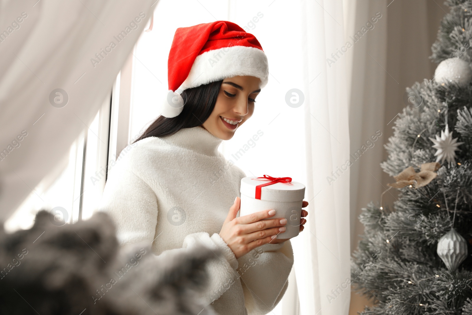 Photo of Woman in Santa hat holding gift box near Christmas tree