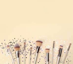 Different makeup brushes and shiny confetti on beige background, flat lay. Space for text