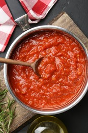 Photo of Homemade tomato sauce in pot, spoon and fresh ingredients on dark table, flat lay