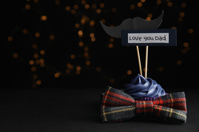 Photo of Composition with cupcake and bow tie on black table against blurred lights, space for text. Happy Father's day