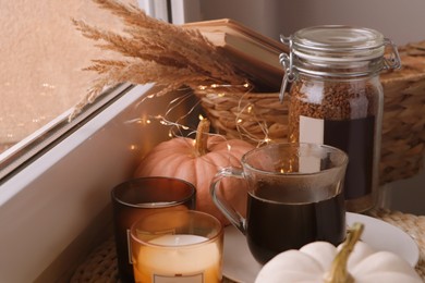 Photo of Cup of hot drink, jar with coffee beans, candles and pumpkins on window sill indoors