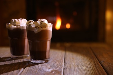 Glasses with hot cocoa and marshmallows on wooden table near fireplace, space for text