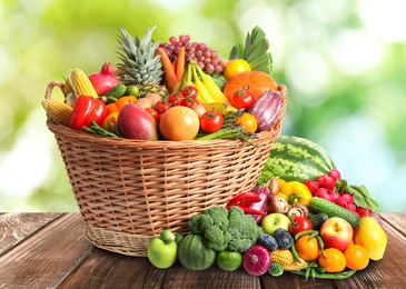 Image of Wicker basket with different fresh organic vegetables and fruits on wooden table against blurred green background 