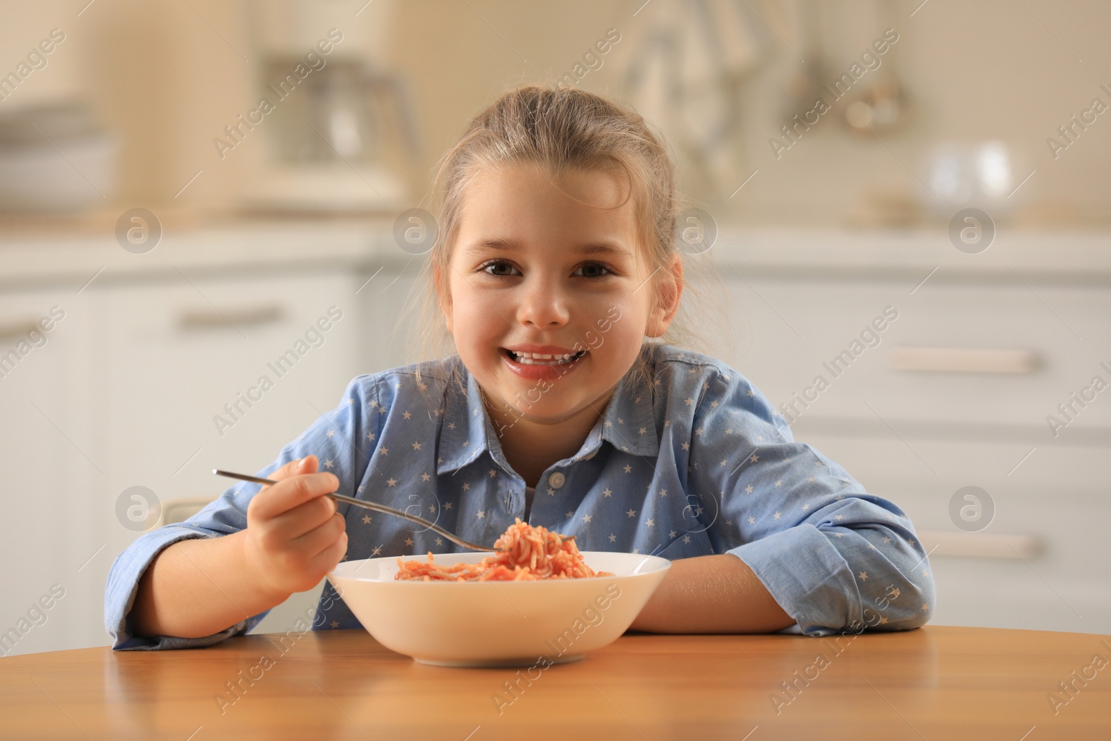 Photo of Cute little girl eating tasty pasta at table in kitchen