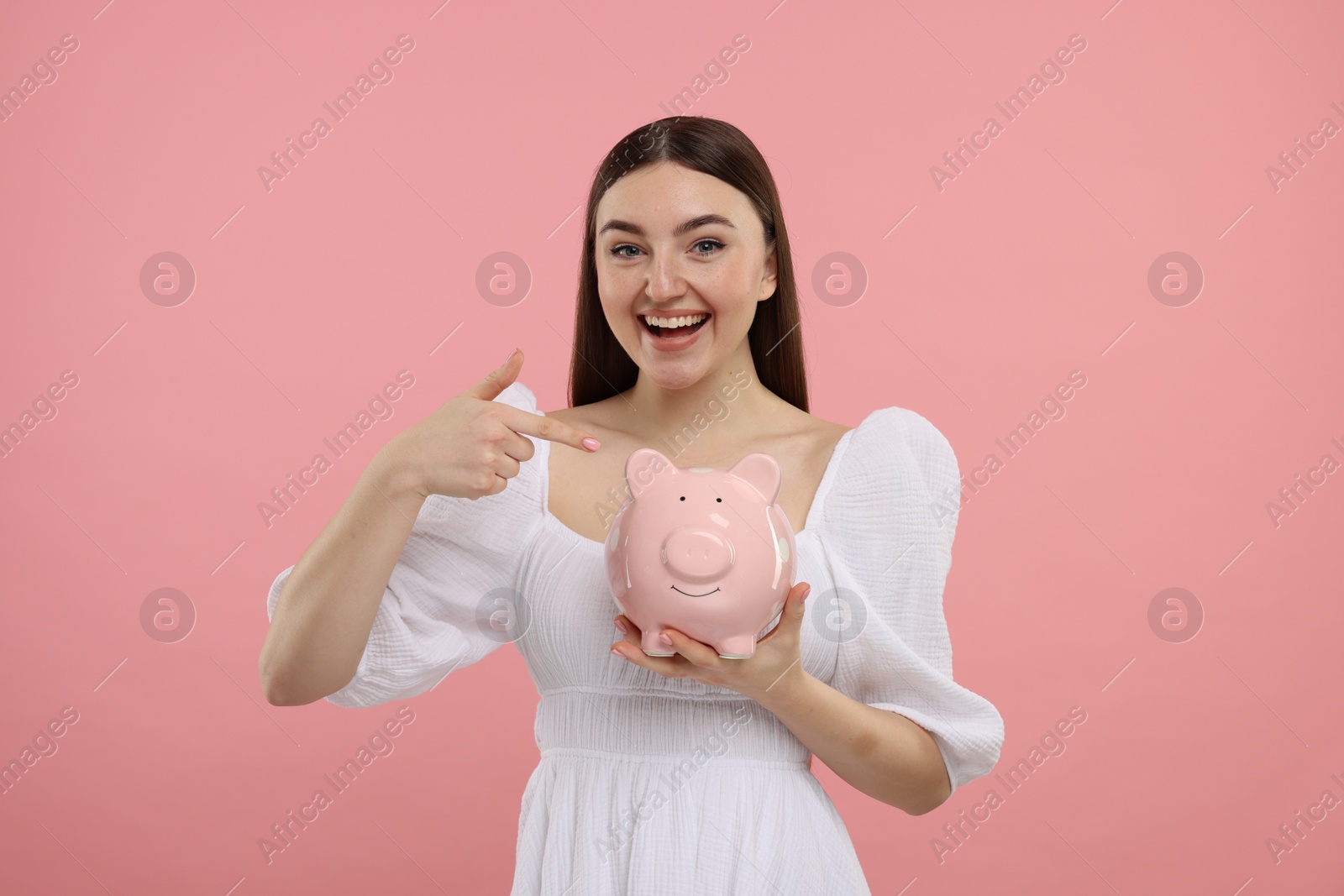 Photo of Happy woman pointing at piggy bank on pink background