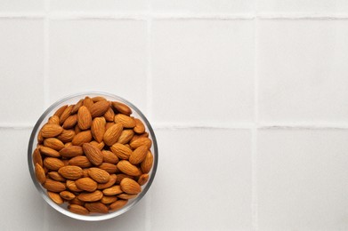 Photo of Bowl of delicious almonds on white tiled table, top view. Space for text