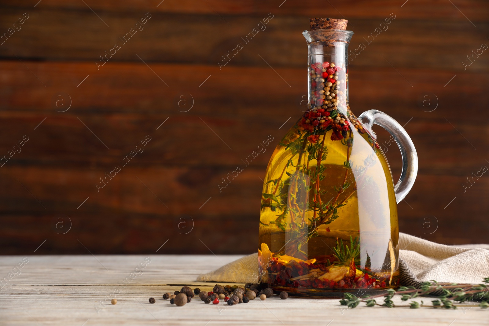 Photo of Glass jug of cooking oil with spices and herbs on white wooden table. Space for text