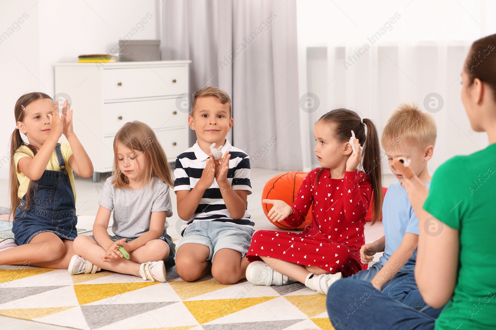 Photo of Nursery teacher and group of cute little children studying with fun on bright rug in kindergarten. Playtime activities