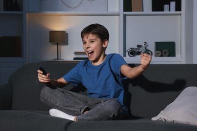 Photo of Emotional boy watching TV and holding remote control on sofa at home