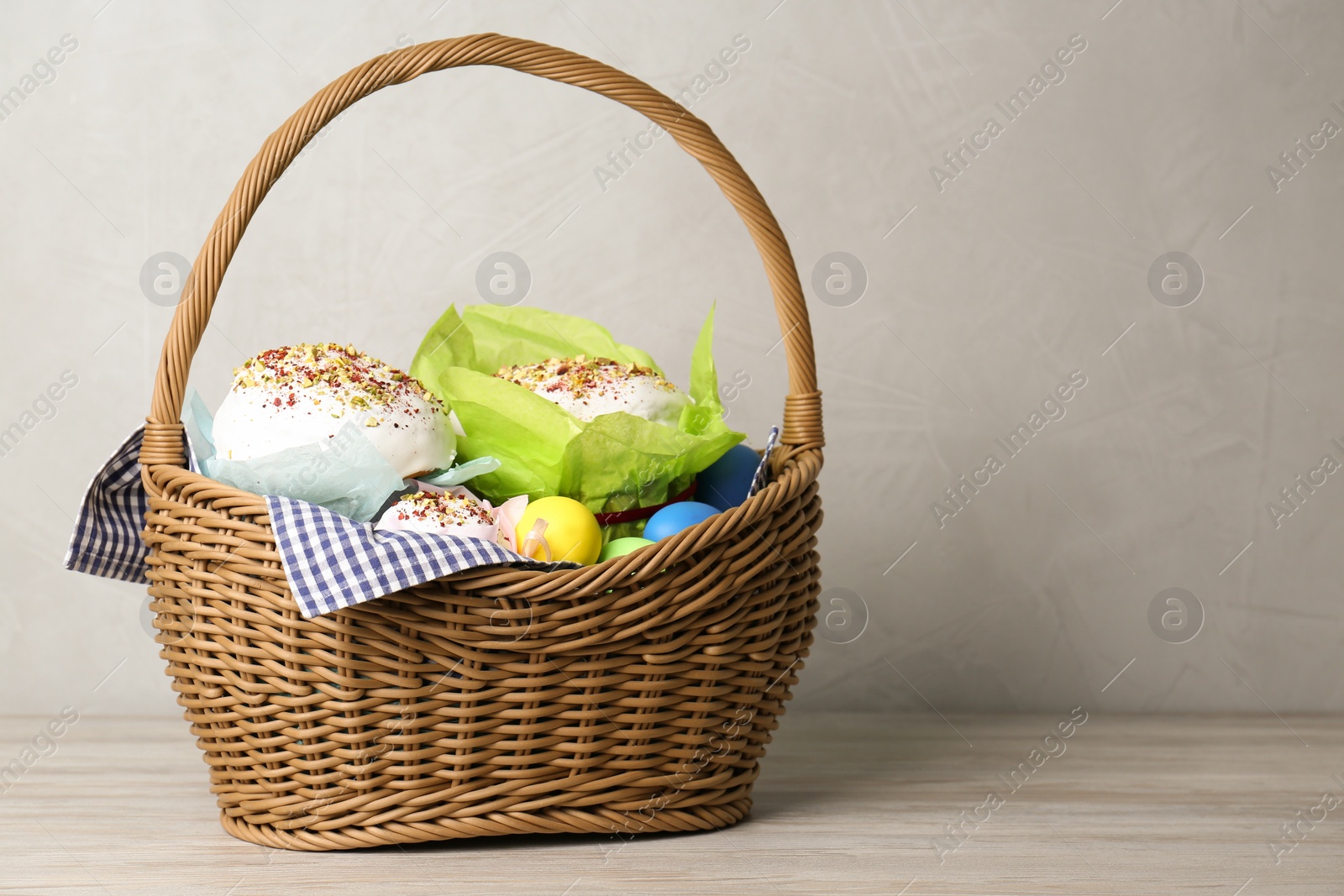 Photo of Basket with delicious Easter cakes and dyed eggs on light wooden table. Space for text