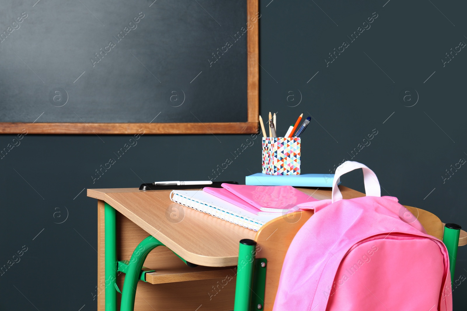 Photo of Wooden school desk with stationery and backpack near blackboard on grey wall