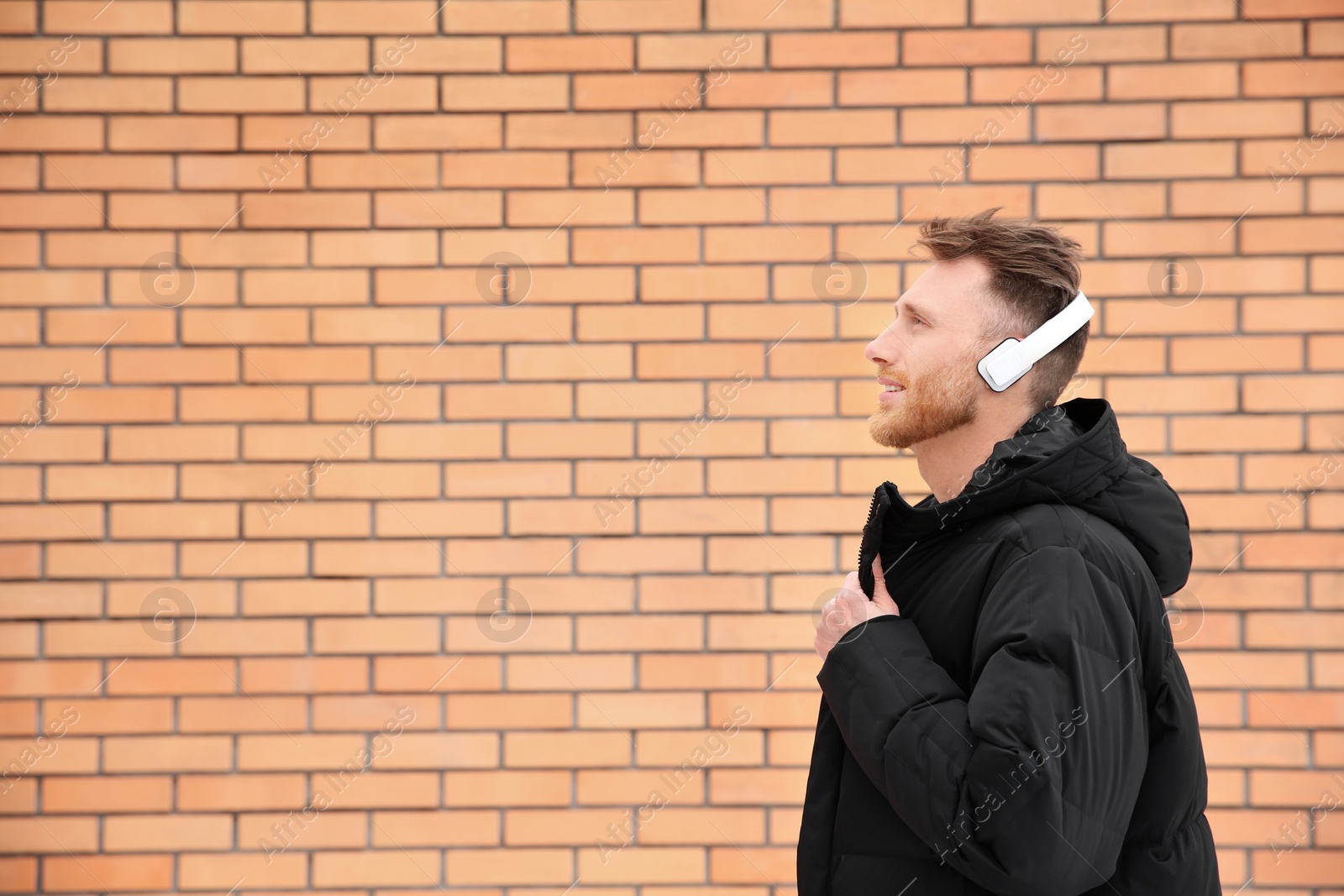 Photo of Young man listening to music with headphones against brick wall. Space for text