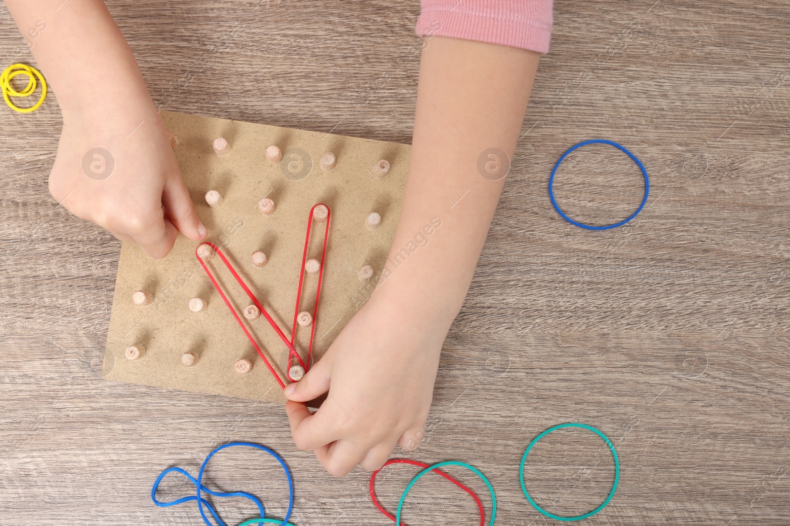 Photo of Motor skills development. Girl playing with geoboard and rubber bands at wooden table, top view