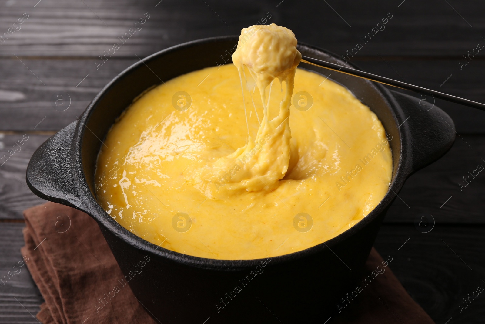 Photo of Dipping piece of ham into fondue pot with melted cheese on black wooden table, closeup