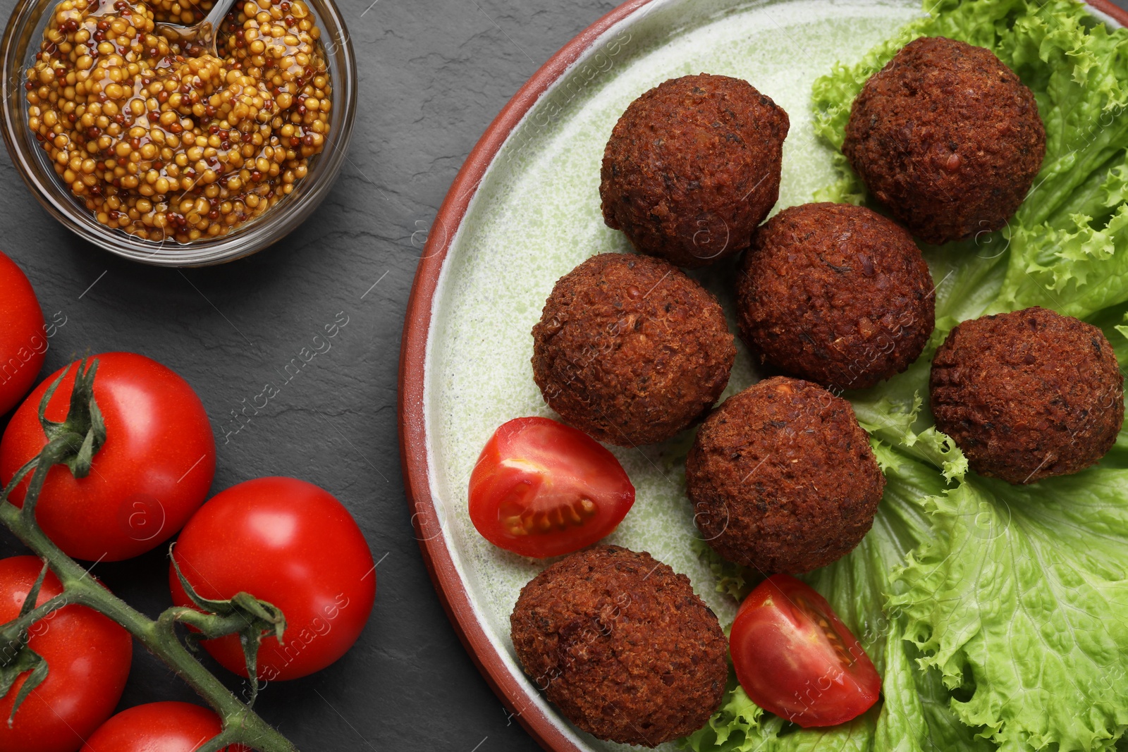 Photo of Delicious falafel balls, tomatoes, lettuce and grain mustard on dark grey textured table, flat lay. Vegan meat products