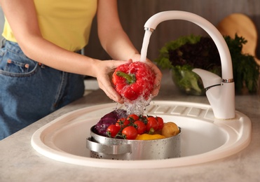 Photo of Woman washing fresh red bell pepper in kitchen sink, closeup