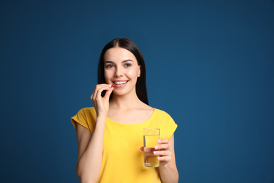 Photo of Young woman with glass of water taking vitamin capsule on blue background. Space for text