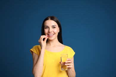 Young woman with glass of water taking vitamin capsule on blue background. Space for text