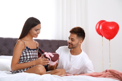 Photo of Young man presenting gift to his girlfriend in bedroom decorated with air balloons. Celebration of Saint Valentine's Day