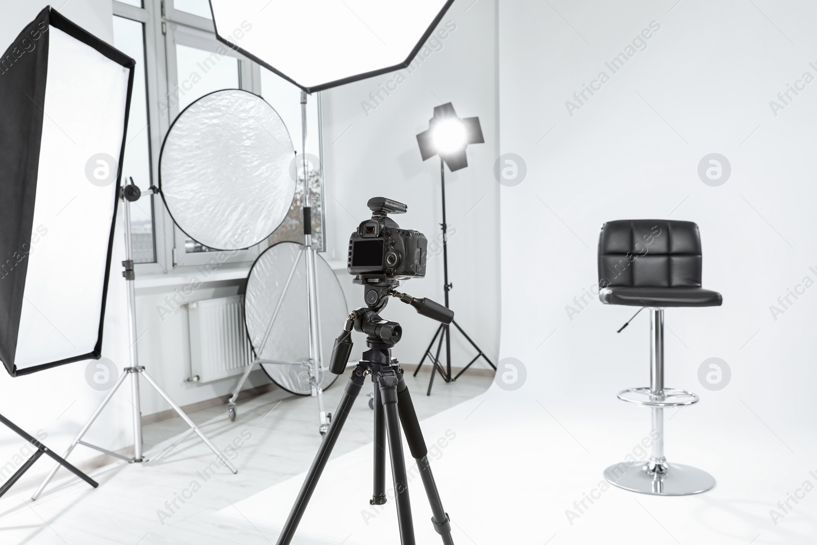 Photo of Tripod with camera, bar stool and professional lighting equipment in modern photo studio