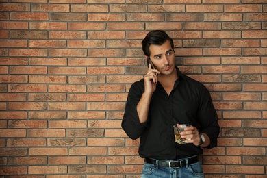 Young man with glass of whiskey talking on phone near brick wall indoors. Space for text
