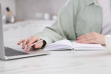 Photo of Woman with notebook working on laptop at white marble table in kitchen, closeup