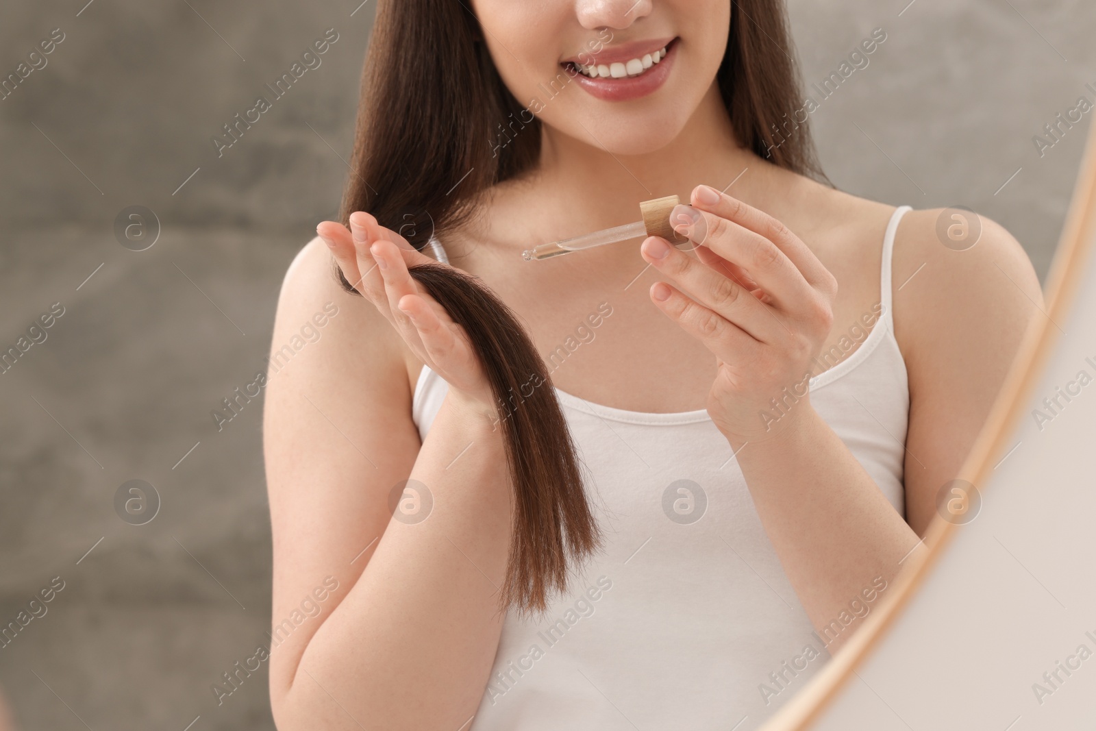 Photo of Happy woman applying essential oil onto hair near mirror, closeup