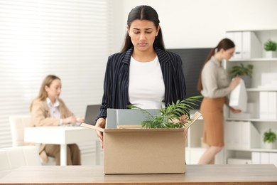 Photo of Unemployment problem. Frustrated woman with box of personal belongings at table in office