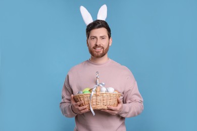 Portrait of happy man in cute bunny ears headband holding wicker basket with Easter eggs on light blue background