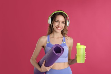Photo of Beautiful woman with headphones, yoga mat and shaker on pink background