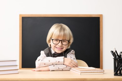 Photo of Happy little school child sitting at desk with books near chalkboard in classroom