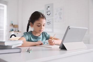 Photo of Little girl doing homework with tablet at table in room