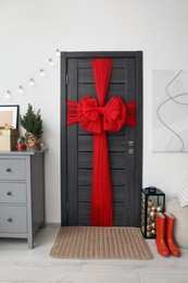 Wooden door with beautiful red bow near chest of drawers and container with baubles indoors. Christmas decoration