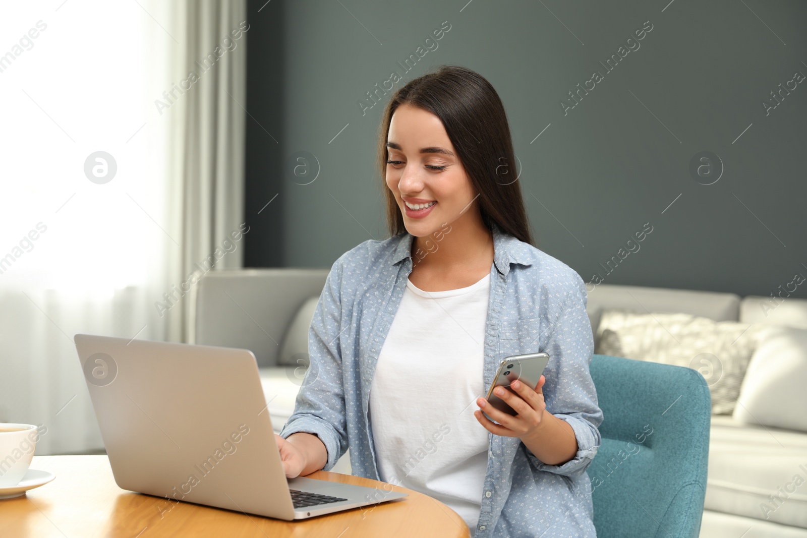Photo of Woman with modern smartphone and laptop at wooden table in living room. Searching information