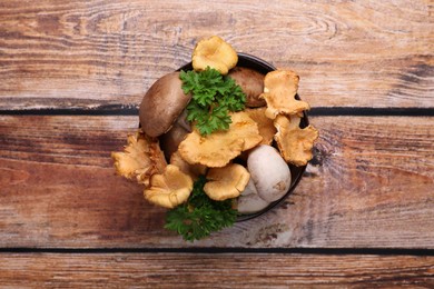 Photo of Bowl of different mushrooms and parsley on wooden table, top view