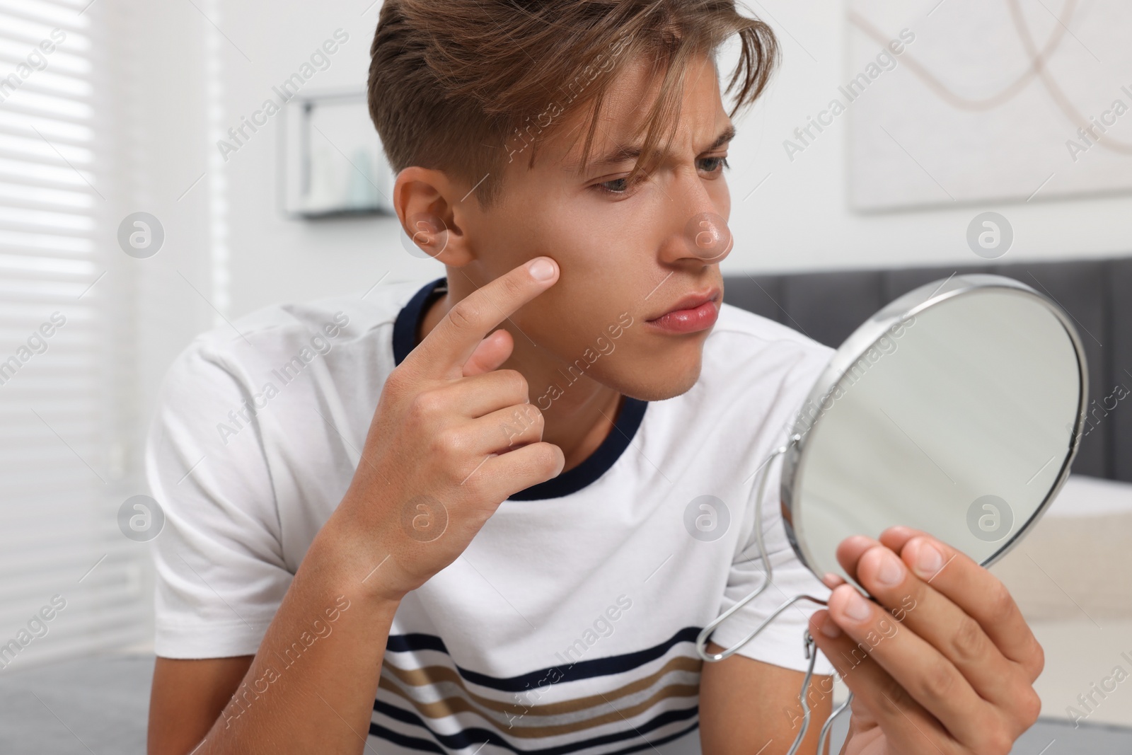 Photo of Upset young man looking at mirror and touching pimple on his face indoors. Acne problem