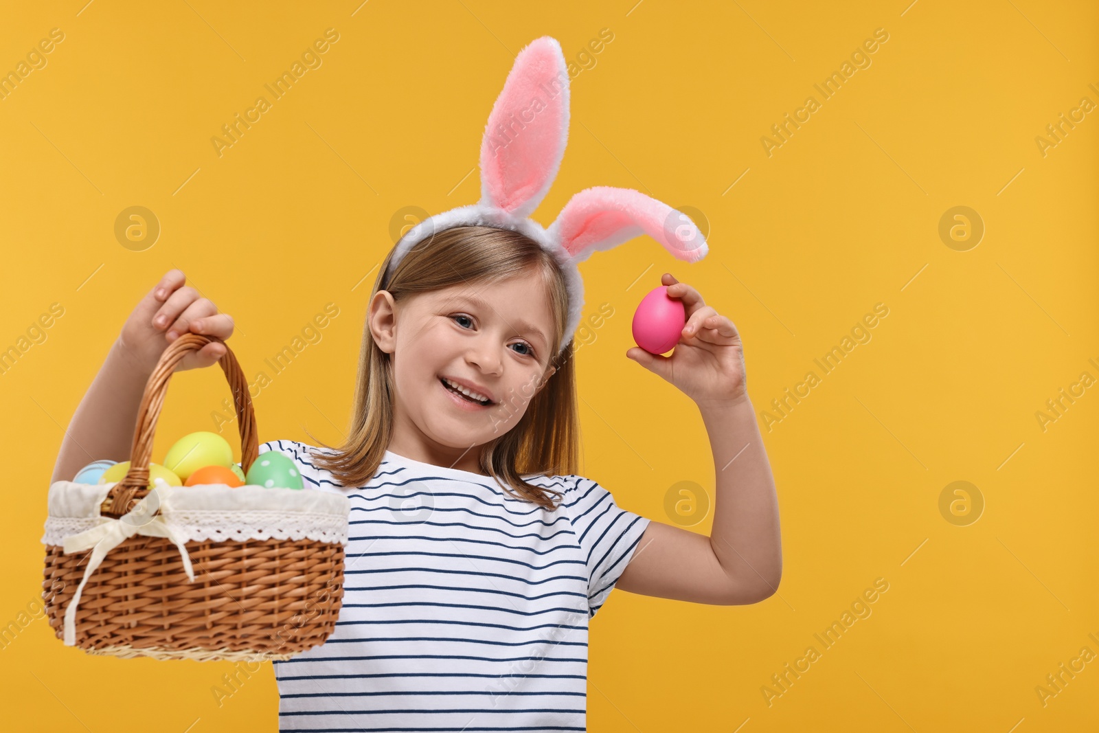 Photo of Easter celebration. Cute girl with bunny ears holding basket of painted eggs on orange background