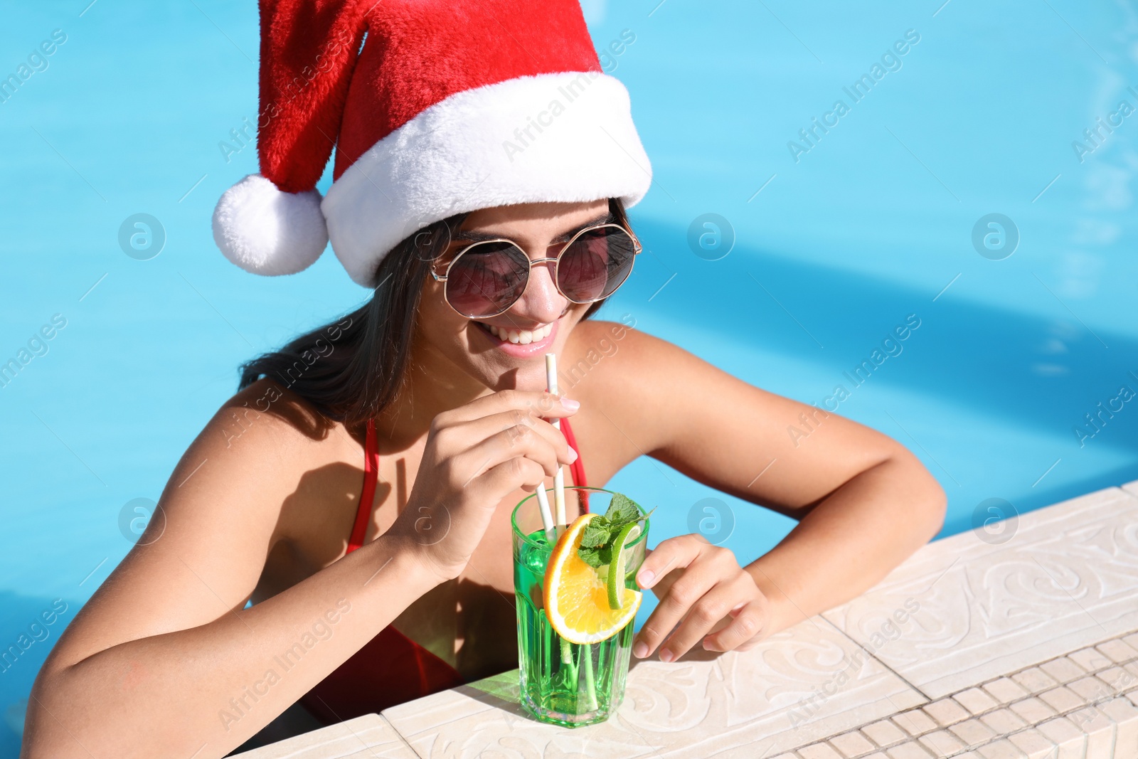 Photo of Young woman wearing Santa Claus hat with refreshing drink in swimming pool. Christmas vacation