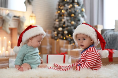 Image of Cute children in Santa hats on floor in room with Christmas tree