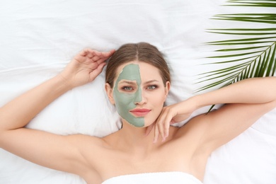 Beautiful woman with clay facial mask and tropical leaf on white fabric, above view