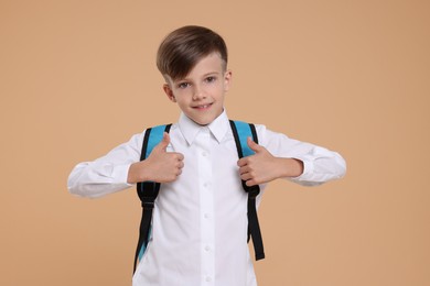 Photo of Cute schoolboy with backpack showing thumbs up on beige background