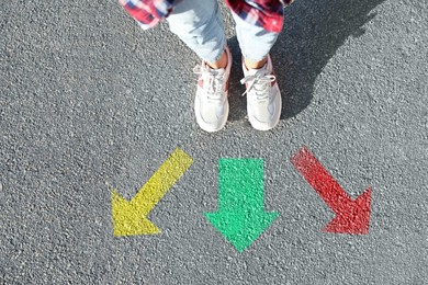 Image of Choice of way. Woman standing in front of drawn marks on road, closeup. Colorful arrows pointing in different directions