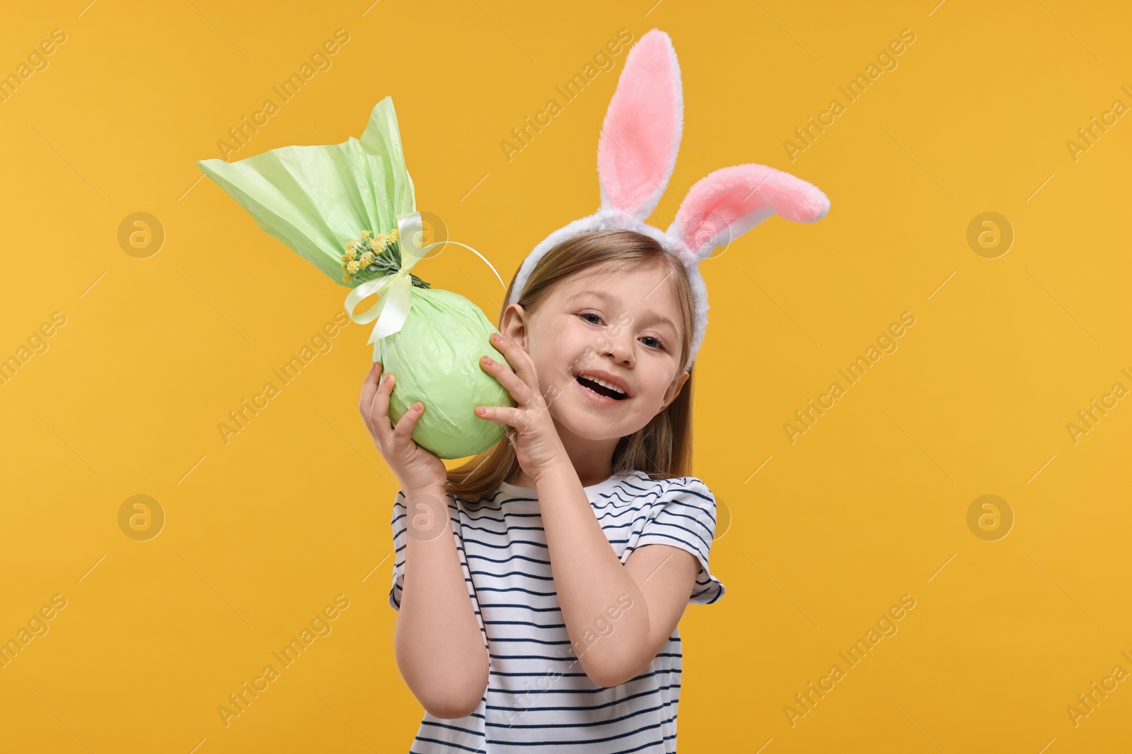 Photo of Easter celebration. Cute girl with bunny ears holding wrapped gift on orange background